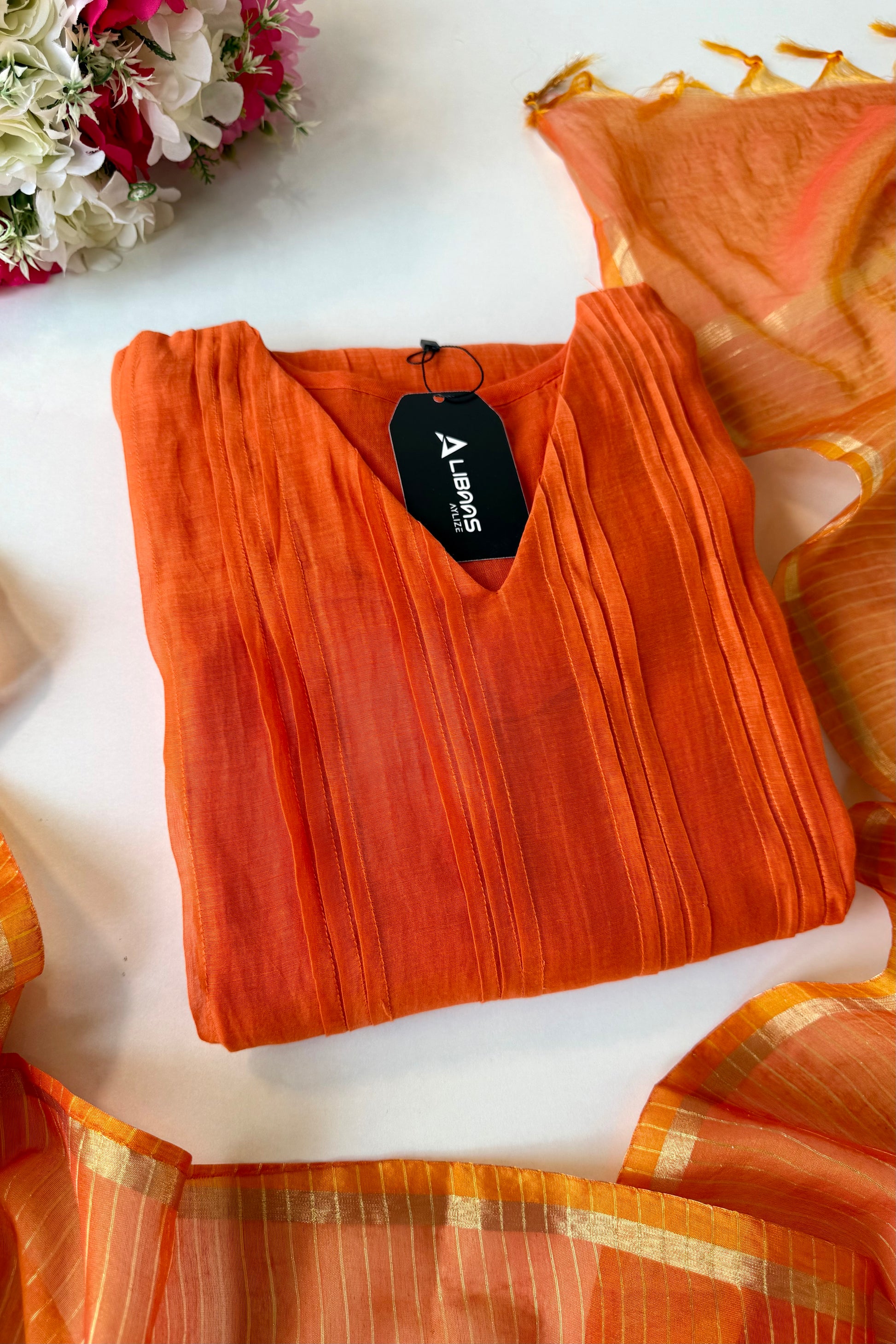 Orange saree with a tag on a white surface, accompanied by a floral arrangement.